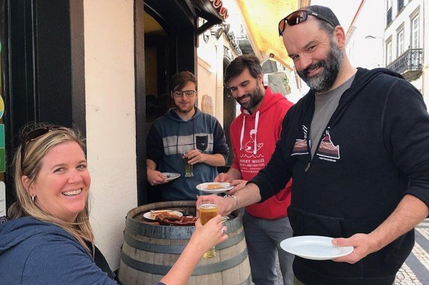 A group of people outdoors, some holding plates and glasses, near a barrel and a building entrance. 2