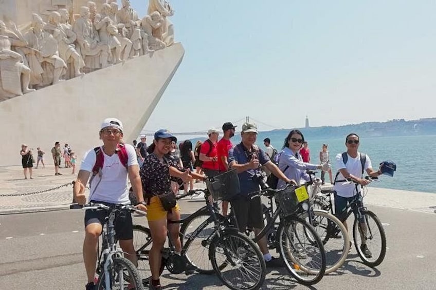 A group of people riding bicycles along a waterfront promenade with sculptures and a bridge in the background. 1