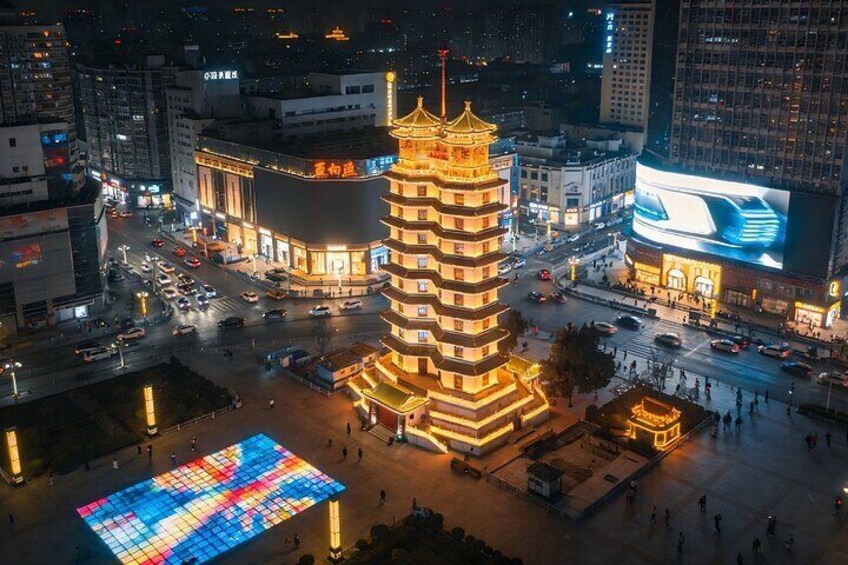 A cityscape at night with a prominent pagoda-like structure in the center, surrounded by illuminated buildings and a large digital billboard. 1