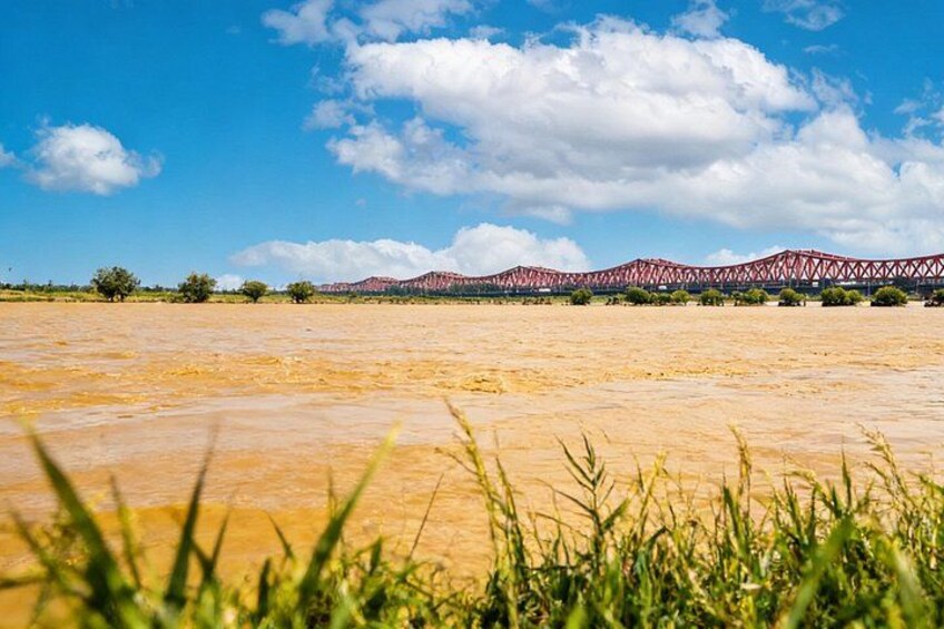 A flat, arid landscape with sparse vegetation in the foreground and a red bridge in the background. 2