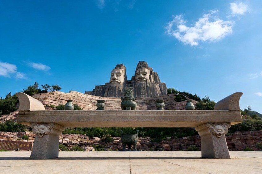 A large stone statue with a face, a bench in the foreground, and a mountainous background. 1