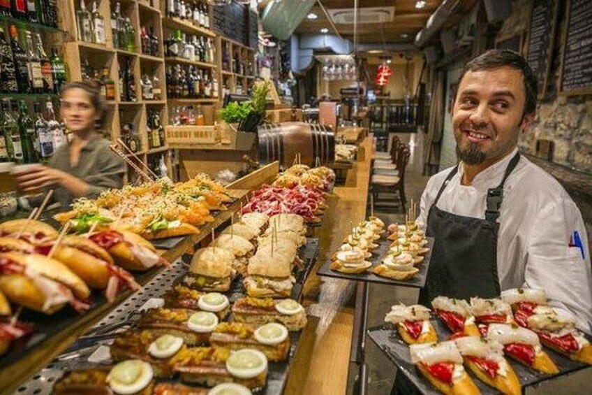 Un puesto de comida con diversos platos, una persona con mandil de chef y estanterías llenas de botellas al fondo. 4