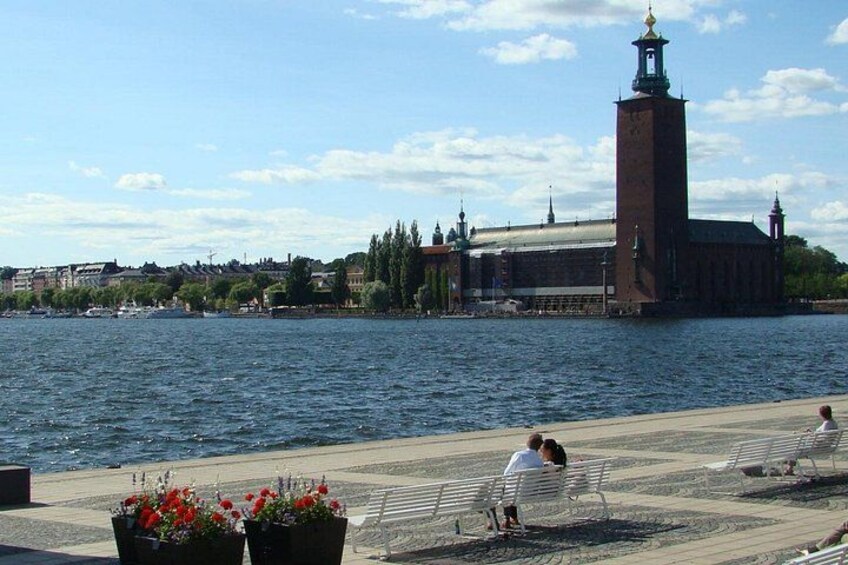 A riverside walkway with benches, a tall brick building with a tower, and a distant cityscape. 1