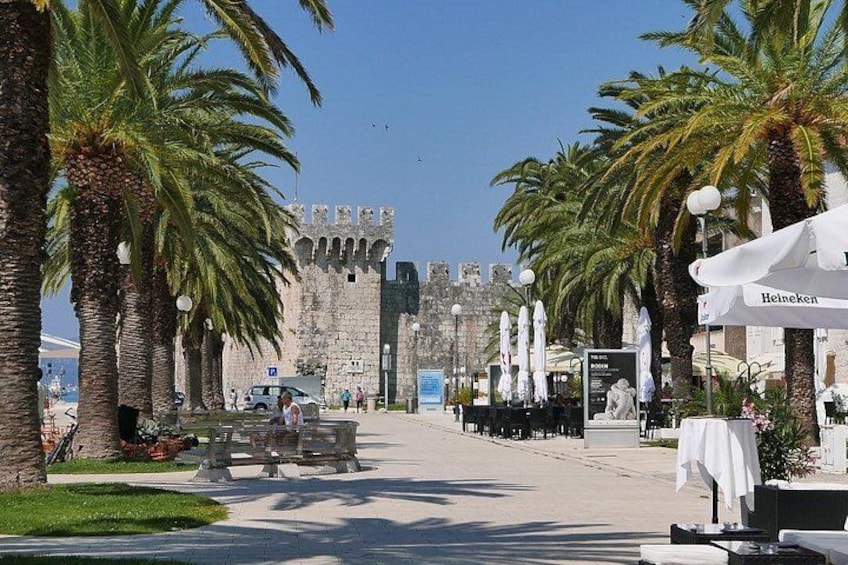 A coastal promenade with palm trees, a historic stone building, and outdoor seating areas. 1