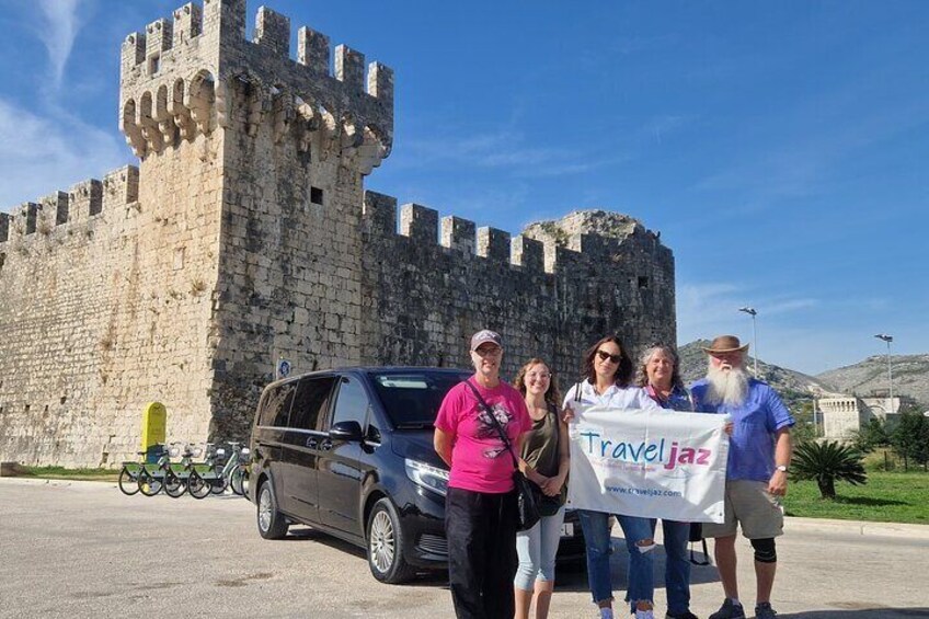A group of people in front of a historic stone castle with a car parked nearby. 4