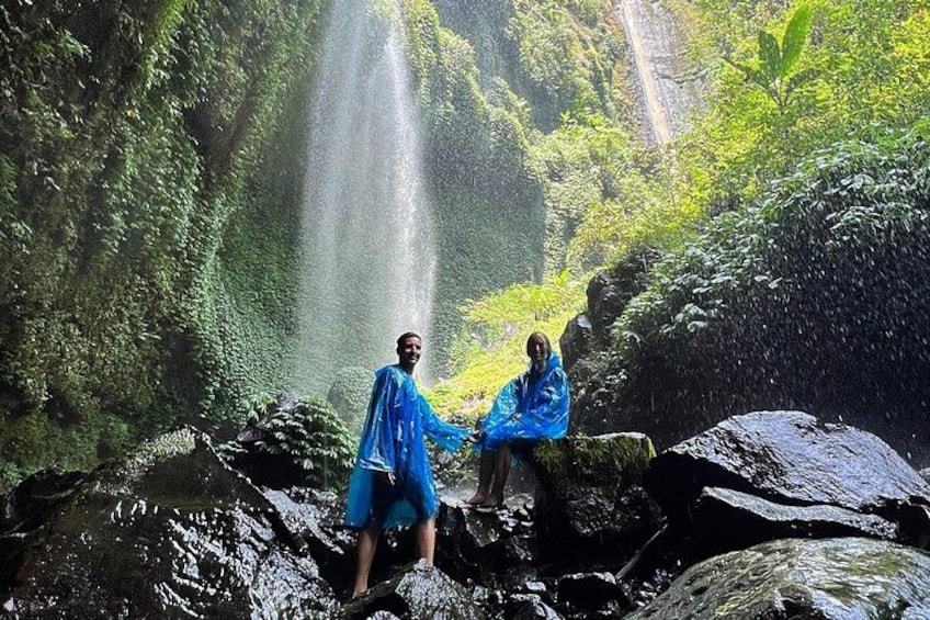 Two people in blue raincoats sitting on rocks near a waterfall in a lush green forest. 3