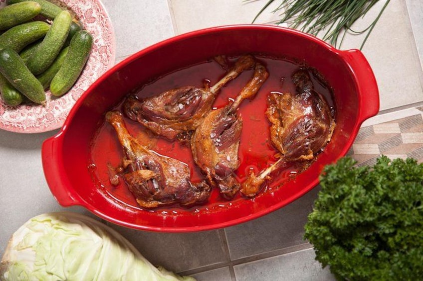 A red baking dish with cooked meat, a bowl of green vegetables, and a cabbage on a tiled surface. 3
