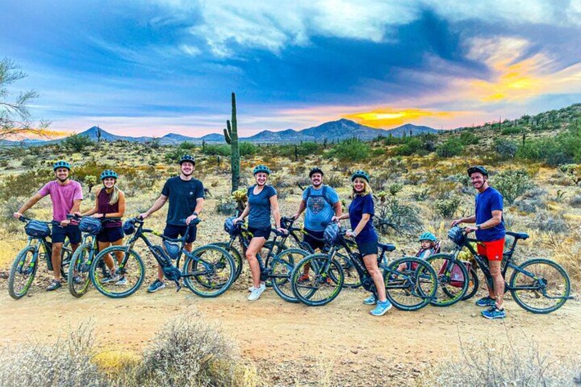 Een groep mensen met fietsen in een woestijnlandschap, met cactussen en bergen op de achtergrond. 3