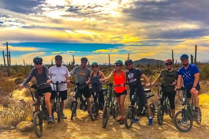 Een groep mensen op fietsen in een woestijnlandschap met schaarse begroeiing en een heldere lucht. 1