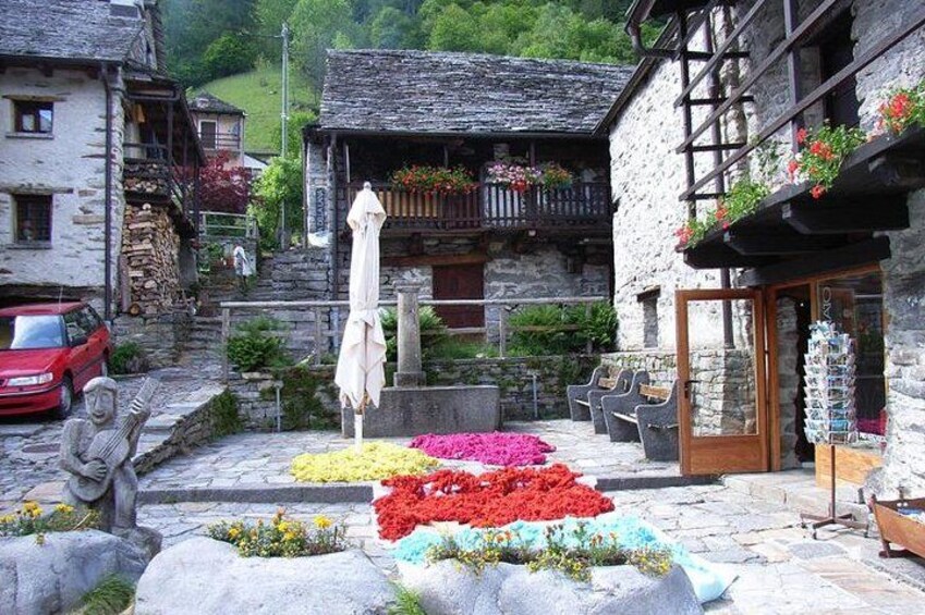A stone building with colorful flower beds in front, a red car parked nearby, and a wooden bench. 5