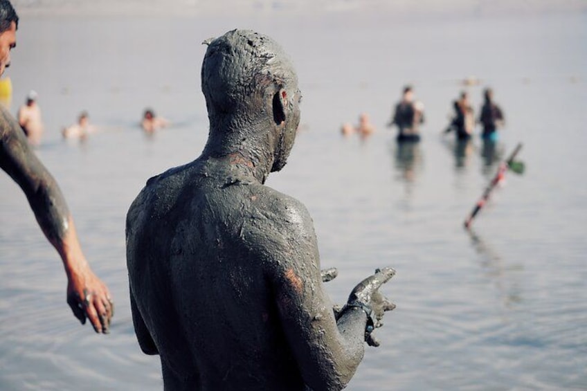 A person covered in mud, standing in water with others swimming in the background. 2