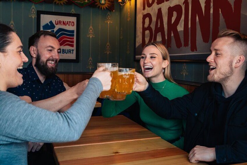 Un groupe de personnes trinquant avec des verres de bière dans un bar où est affiché un panneau sur lequel on peut lire : « UR NATO BURT ». 2