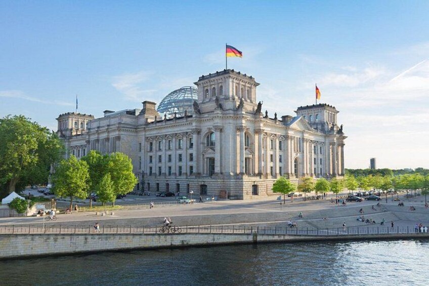 Ein großes, historisches Gebäude mit Flaggen, einer Uferpromenade und einem gepflasterten Platz mit Bäumen. 1