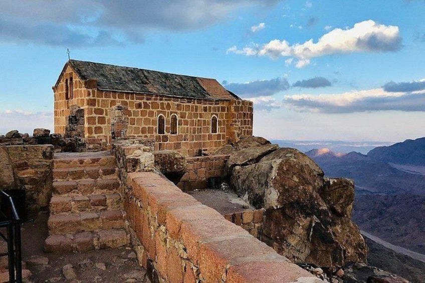 A stone building with a sloped roof, surrounded by a stone wall and steps leading up to it. 2