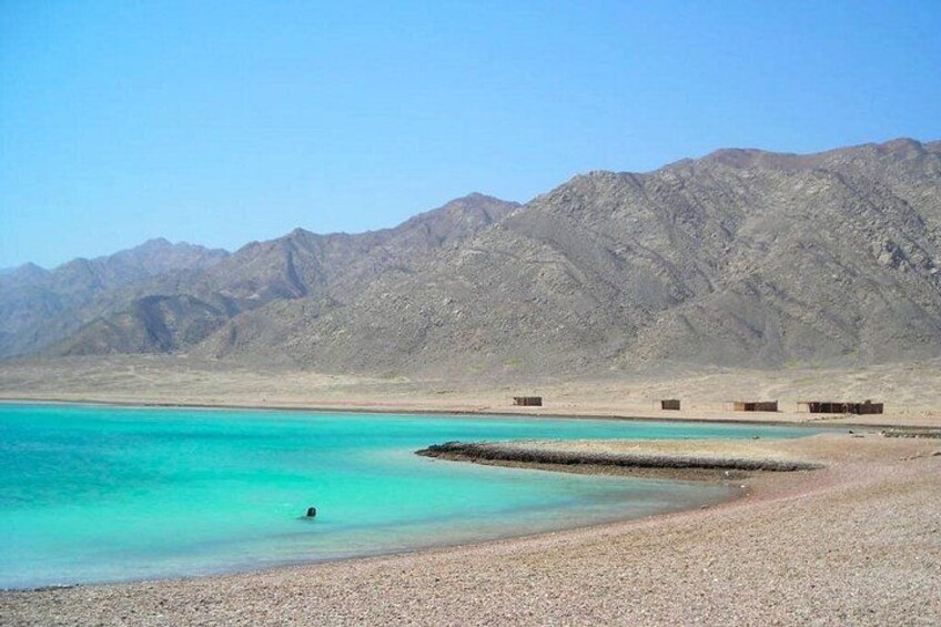 A beach with turquoise water, a person swimming, and mountains in the background. 3