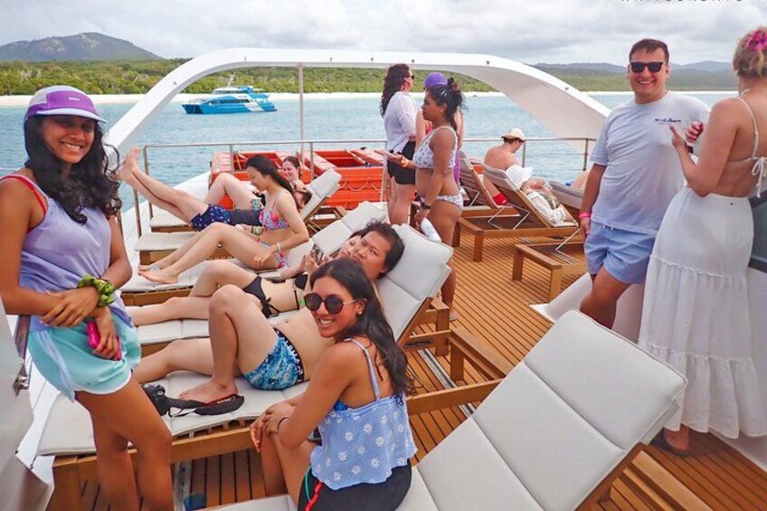 A group of people on a boat with wooden deck and white lounge chairs, surrounded by water and a distant island. 5
