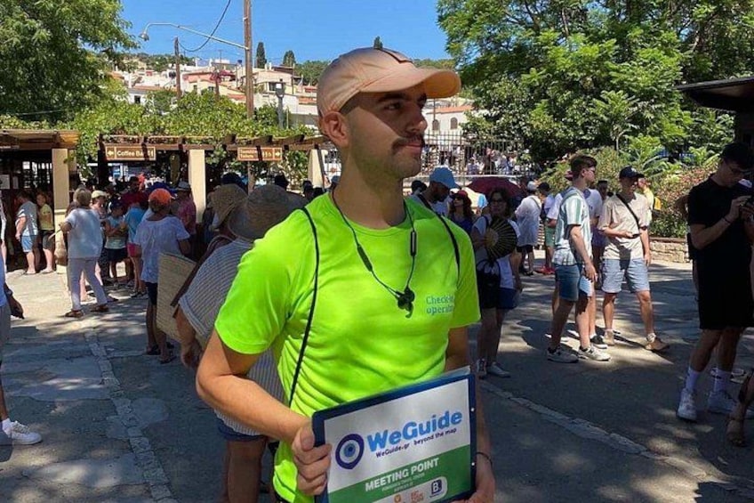 Una persona sostiene un cartel de WeGuide en un entorno exterior concurrido, con edificios y árboles de fondo. 5