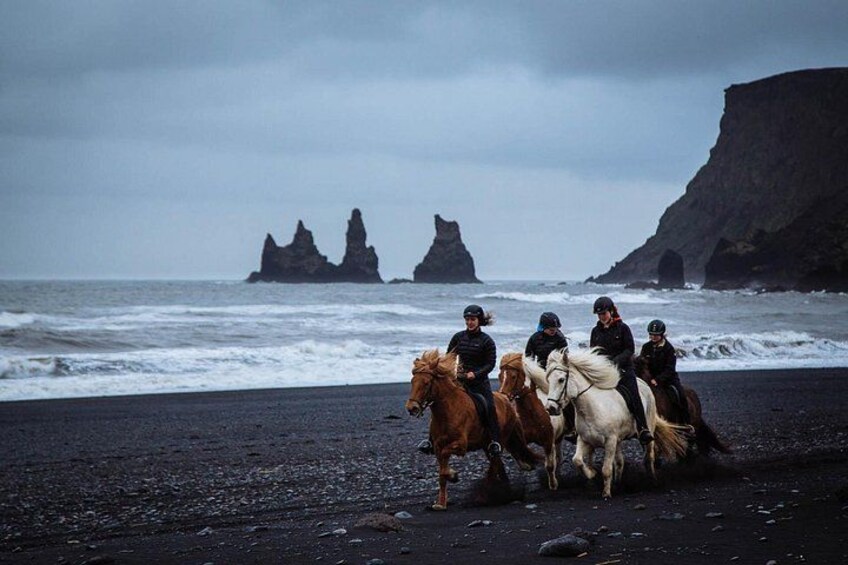 A group of people riding horses on a black sand beach with rocky formations in the background. 1