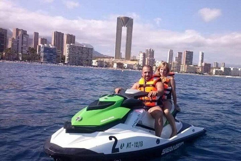 A jet ski with two people wearing life jackets on a body of water with a city skyline in the background. 4