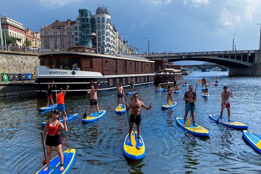 Menschen stehen auf Paddleboards auf einem Fluss, im Hintergrund sind Gebäude und eine Brücke zu sehen. 1