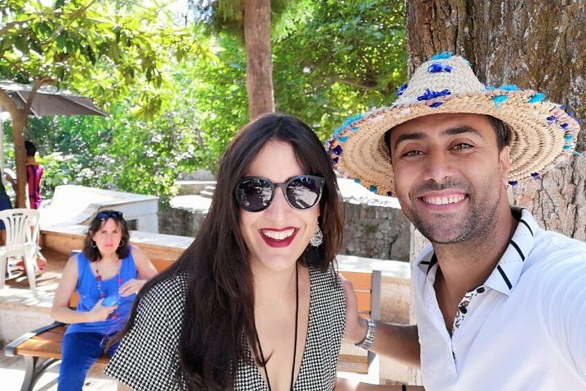 A person wearing a straw hat with blue flowers, a person in a black and white patterned top, and a person in a blue top sitting at a table in a sunny outdoor setting. 5