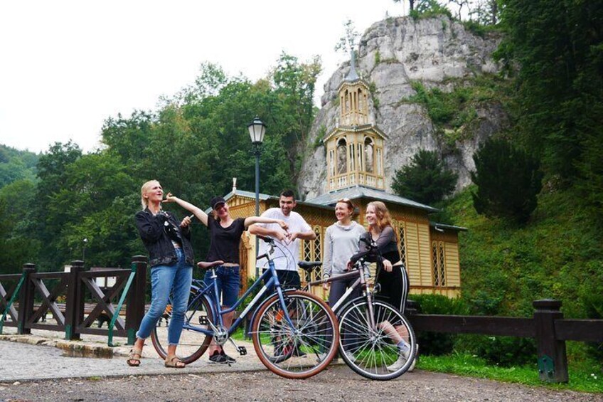 A group of people standing by bicycles in front of a historic building with a bell tower, surrounded by greenery and a rocky landscape. 1