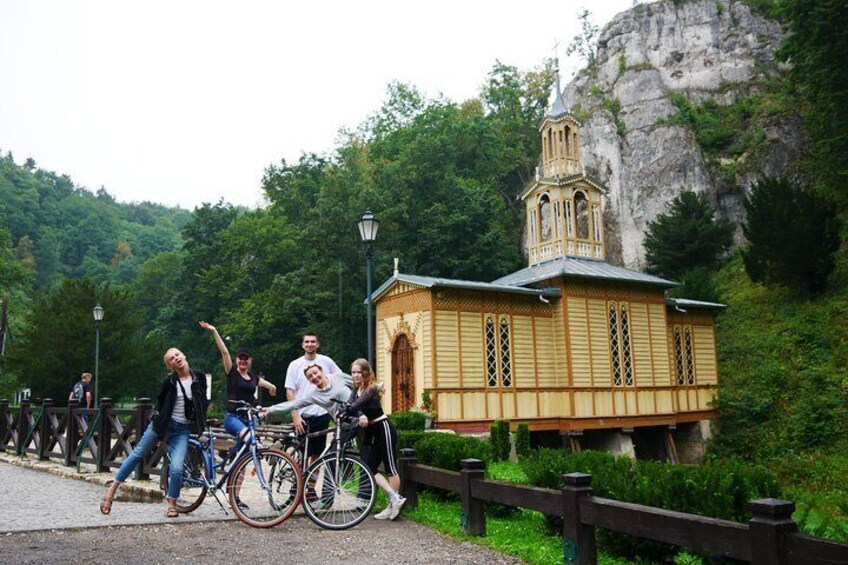 A group of people on bicycles near a small wooden structure with a bell tower, surrounded by lush greenery and a rocky hill. 4