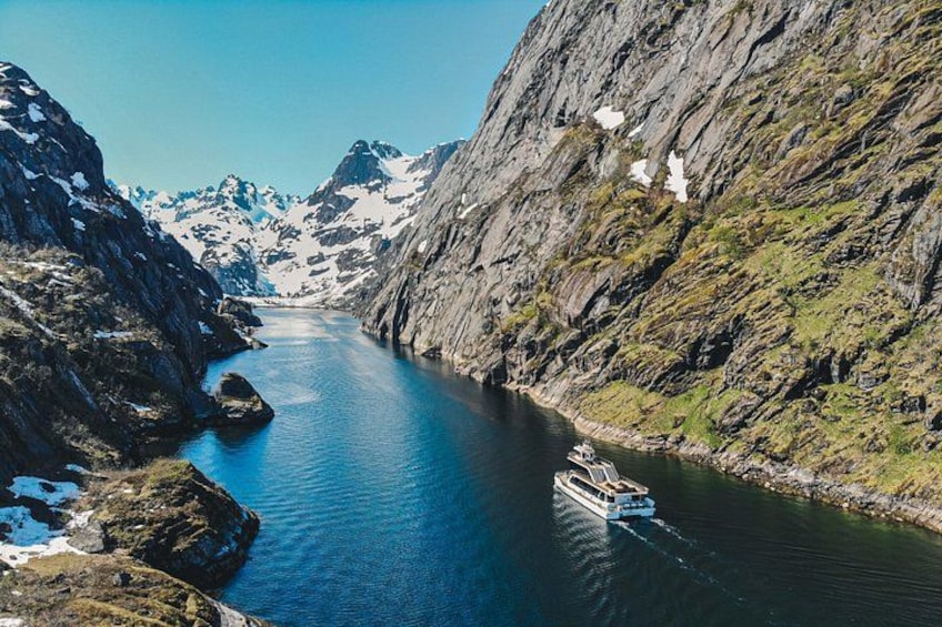 A boat on a narrow fjord surrounded by steep cliffs with patches of snow. 1