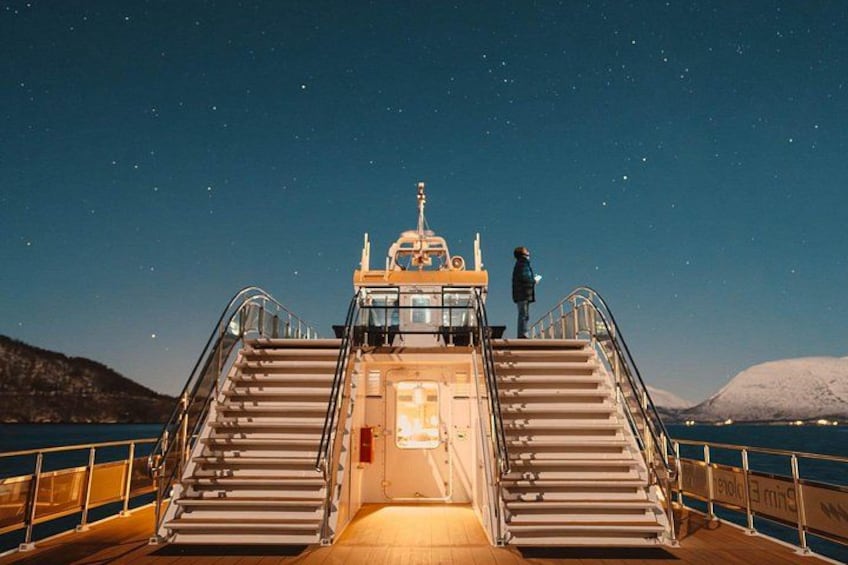 A ship's deck with a person standing at the top of the stairs, looking out at the stars. 4
