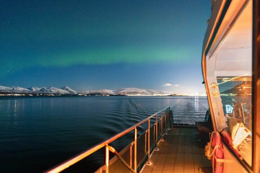 A nighttime view from a boat with the Northern Lights in the sky, calm water, and snow-covered mountains in the distance. 1