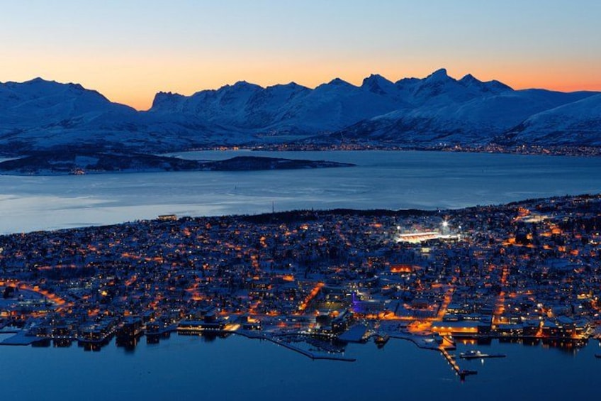 A coastal city at dusk with illuminated buildings, a harbor, and snow-capped mountains in the background. 2