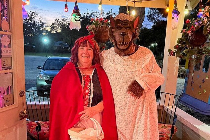 A person in a red cape and another in a wolf costume standing on a porch decorated with lights and plants. 2
