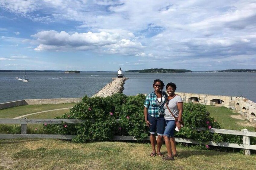 Two people standing by a waterfront with a lighthouse in the background. 3