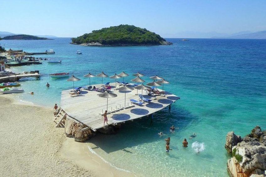 Una playa con un muelle de madera, agua turquesa cristalina y una pequeña isla al fondo. 3