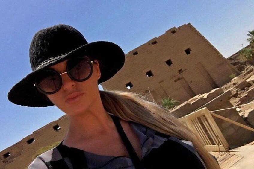 A person wearing a black hat and sunglasses, standing in front of a large, unfinished brick building with multiple small windows. 4