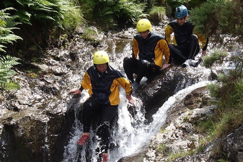 Drie mensen in gele helmen en zwarte neopreenpakken zitten op een rotsachtige richel naast een waterval. 1