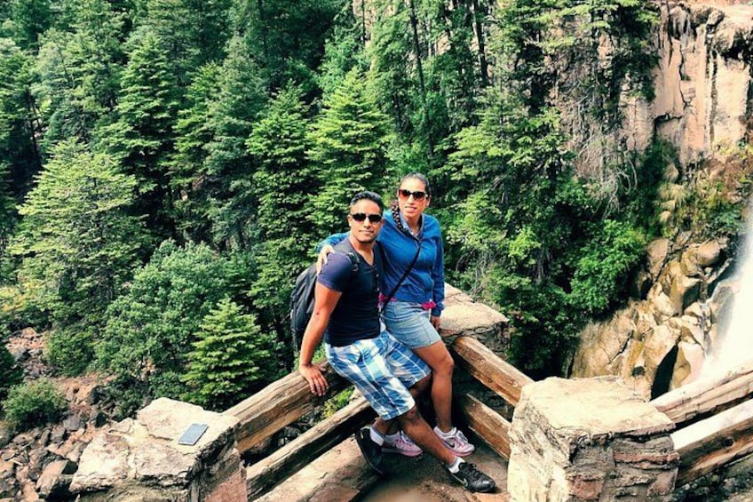 Deux personnes sont assises sur un pont en bois, avec une forêt et une cascade en arrière-plan. 3