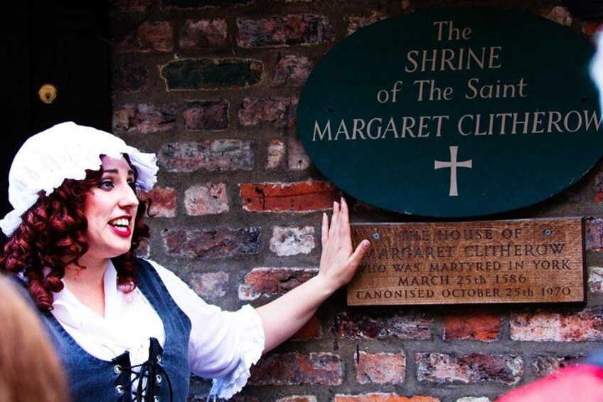 En person i historisk kostume står ved et skilt, der viser 'The Shrine of The Saint Margaret Clitherow' med et kors. 1
