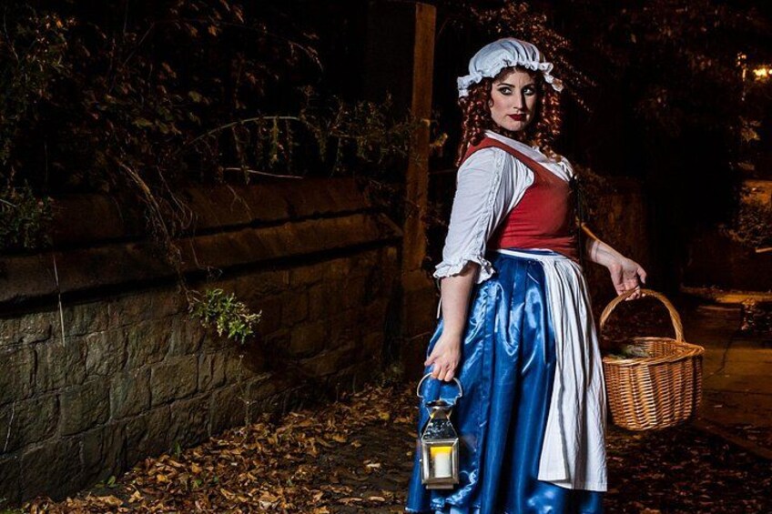 Una persona vestida con traje tradicional sostiene un farol y una cesta, de pie junto a un muro de piedra por la noche. 2