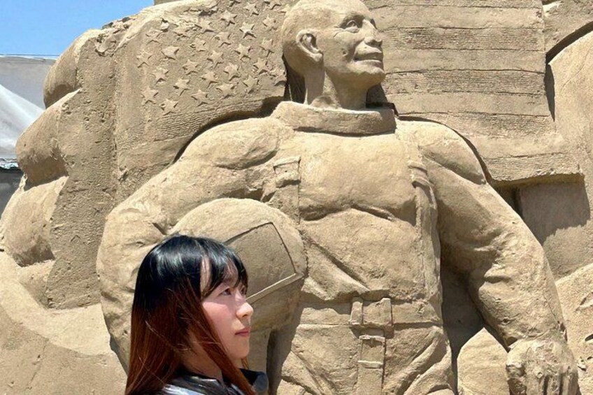A large sand sculpture of a muscular figure with stars on its chest, set against a clear sky. 4