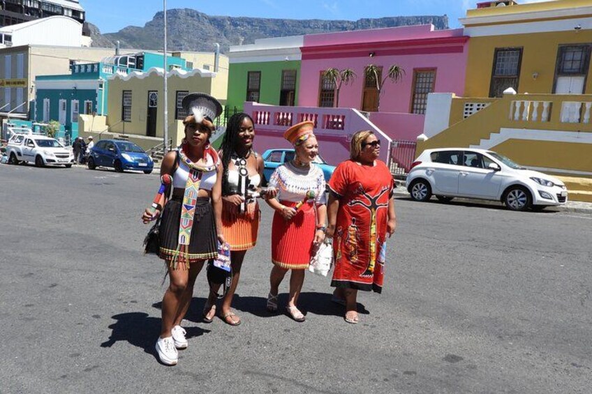 Four people in traditional attire standing in a street with colorful buildings in the background. 4