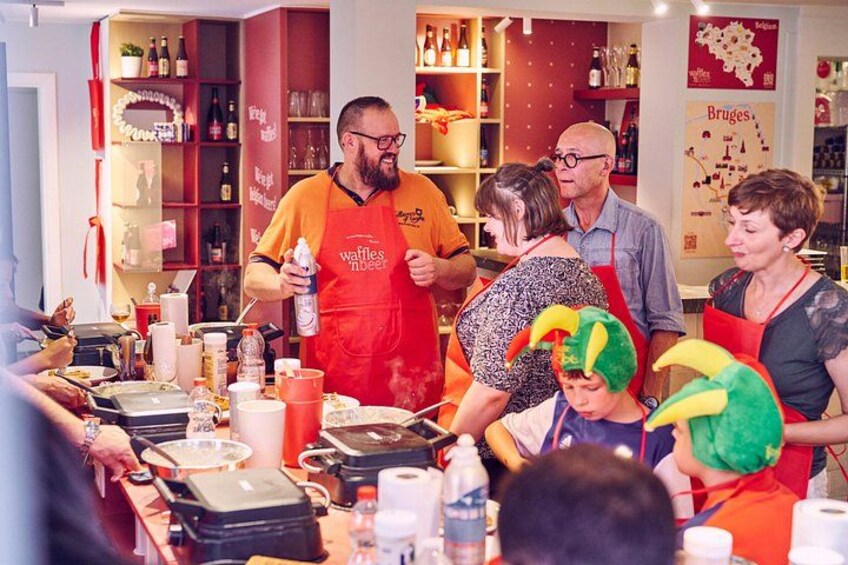 Un groupe de personnes se trouve dans une cuisine, certaines portent des tabliers, et on peut voir des ustensiles de cuisine et des bouteilles sur le plan de travail. 2