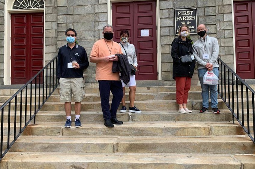 Un gruppo di persone in piedi sui gradini di pietra di fronte a un edificio con porte rosse e un cartello che riporta la scritta 'FIRST PARISH'. 4