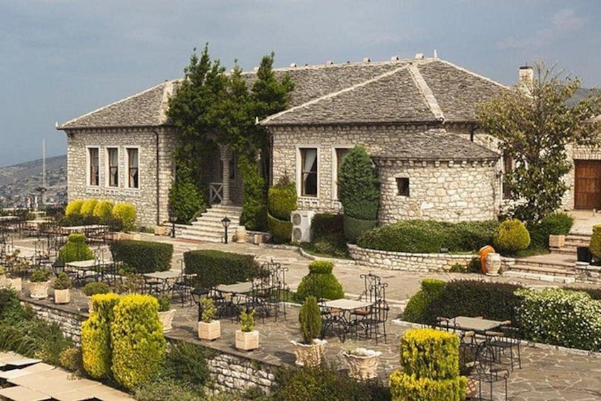 Un bâtiment en pierre avec un toit en tuiles, entouré d’un jardin aménagé avec des arbustes soigneusement taillés et d’une terrasse dallée équipée de mobilier d’extérieur. 1