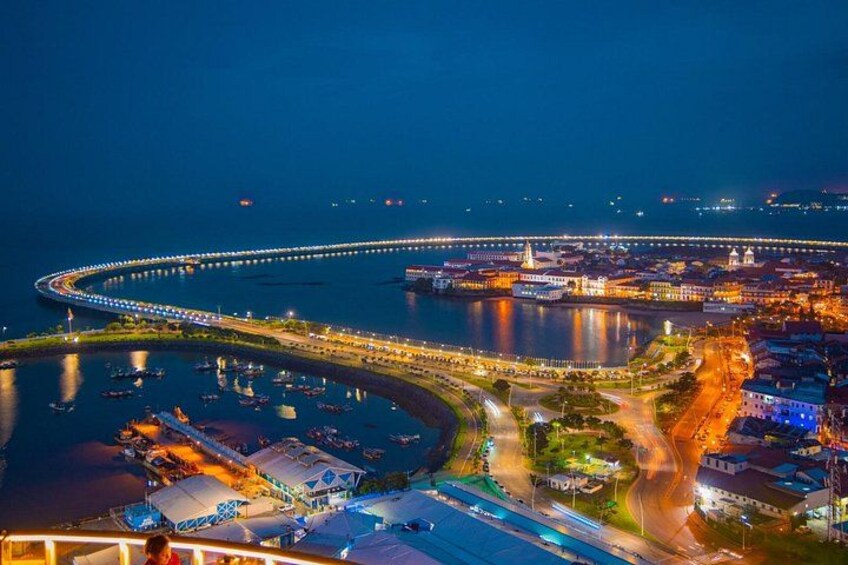 Une vue nocturne d’une ville côtière, avec un pont incurvé imposant, des bâtiments illuminés et une marina remplie de bateaux. 1