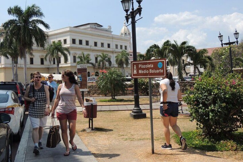 A street scene with pedestrians, a sign indicating 'Plazaoleo Medio Baluarte', palm trees, and a building in the background. 2