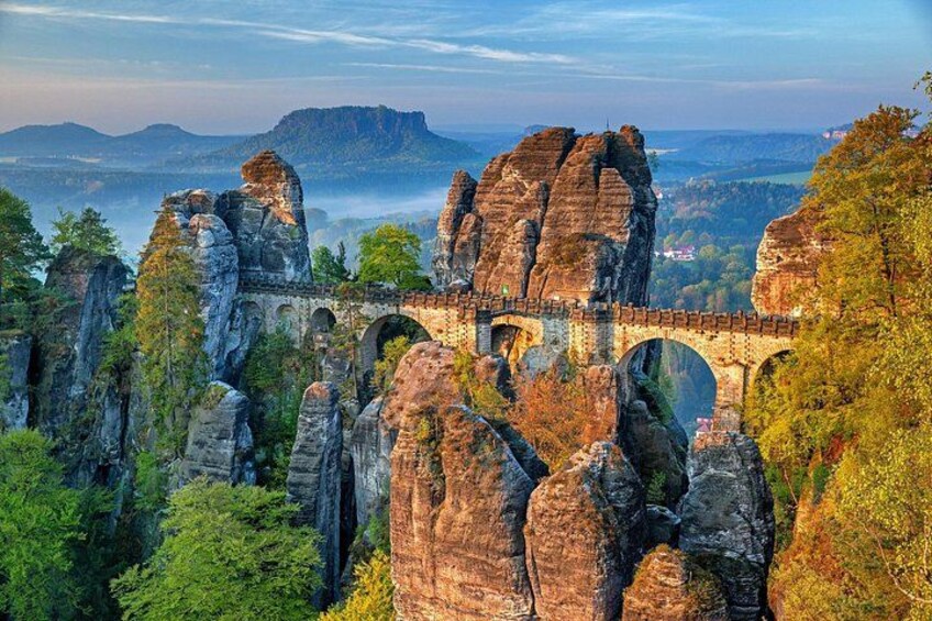 A stone bridge over a rocky gorge with a forested landscape and distant mountains. 1