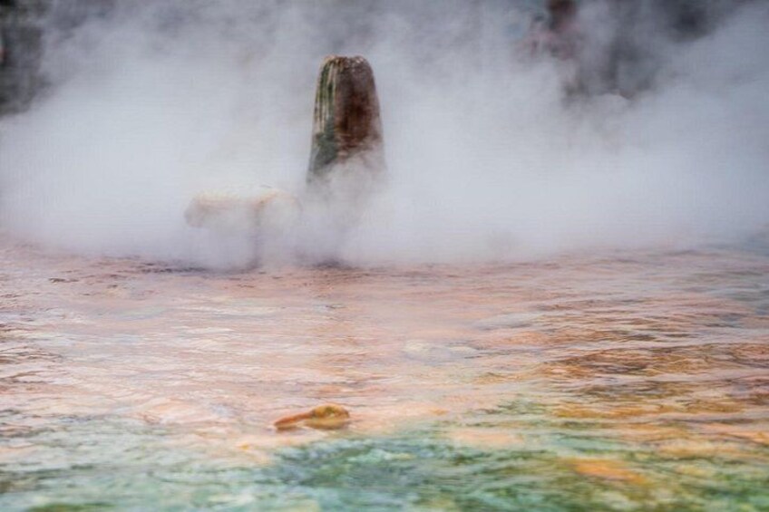 A geothermal area with steam rising from the ground, a rock formation, and a pool of water. 4