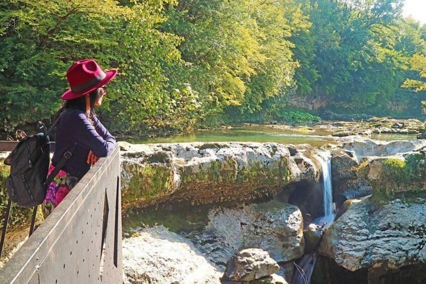 Eine Person mit roter Mütze und violetter Jacke steht auf einer Brücke und blickt auf einen Wasserfall in bewaldeter Landschaft. 4
