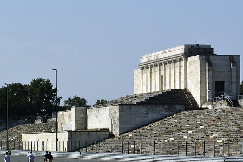 A large, white, classical-style building with steps leading up to it, surrounded by a fence and a few people walking nearby. 3